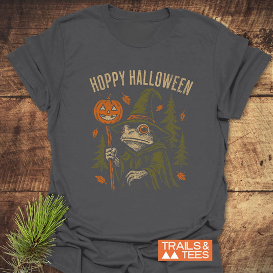 Hoppy Halloween T-Shirt with a frog in a witch’s hat and cloak, holding a jack-o’-lantern staff. Features durable print, soft cotton, and Trails & Tees logo.