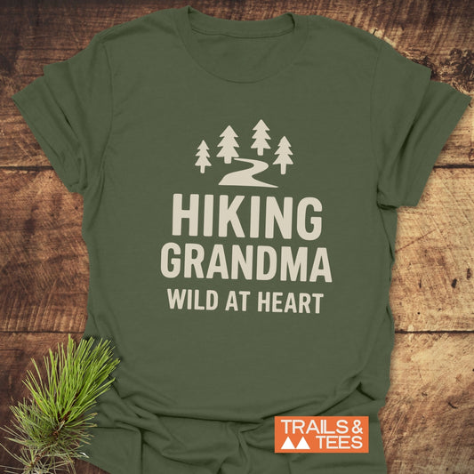 Olive-green tee with pine trees and Hiking Grandma Wild At Heart text, highlighting outdoor adventure spirit. Includes brand TRAILS & TEES and evergreen sprig accent.