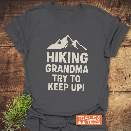 Dark-grey Hiking Grandma - Try To Keep Up T-shirt with mountain print, orange logo, on wood surface with pine sprig; soft, durable, adventure-ready design.