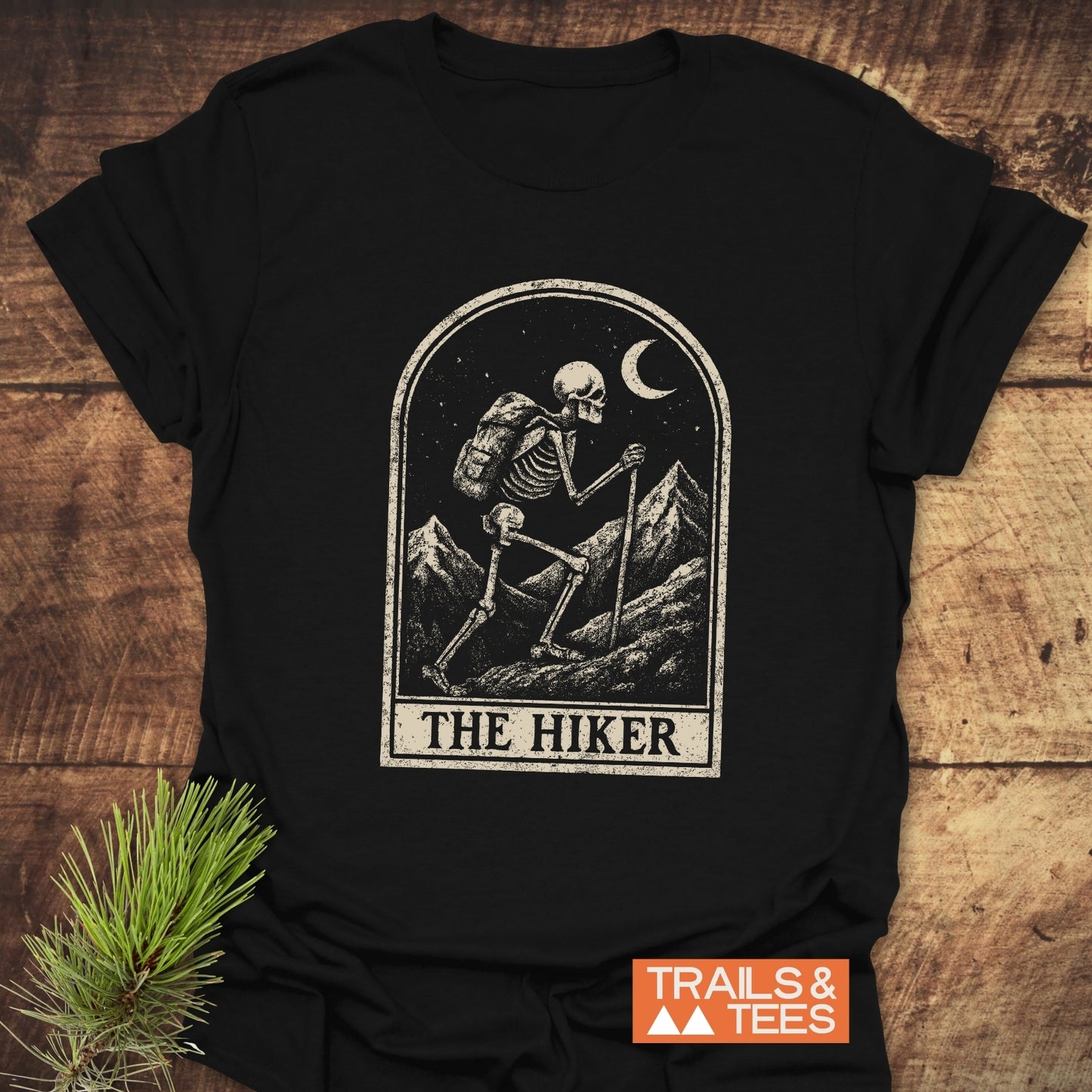 The Hiker Tarot T-Shirt features a distressed skeleton hiking graphic under a crescent moon, with TRAILS & TEES logo, crafted for outdoor enthusiasts.