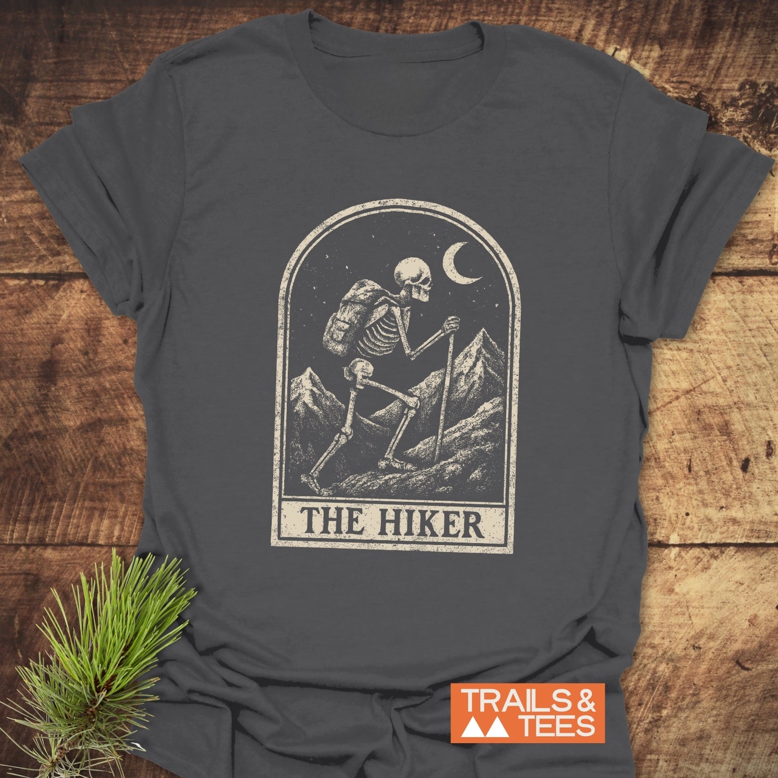 The Hiker Tarot T-Shirt features a skeleton hiking graphic under a crescent moon, with TRAILS & TEES tag, highlighting adventure-ready, soft cotton construction.