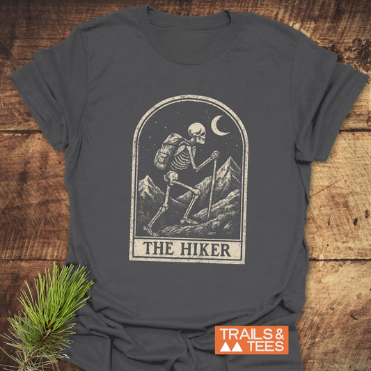 The Hiker Tarot T-Shirt features a skeleton hiking graphic under a crescent moon, with TRAILS & TEES tag, highlighting adventure-ready, soft cotton construction.