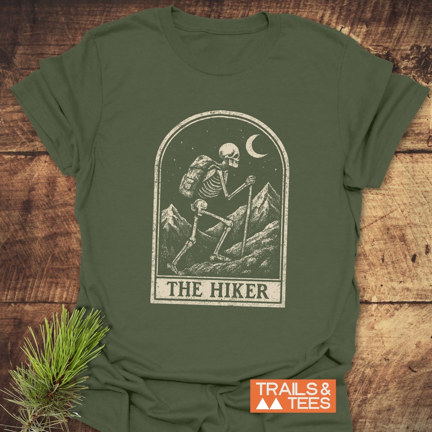 The Hiker Tarot T-Shirt flat-lay with a skeleton hiker illustration, crescent moon, mountain peaks; features durable print, soft cotton, pre-shrunk for comfort and fit.