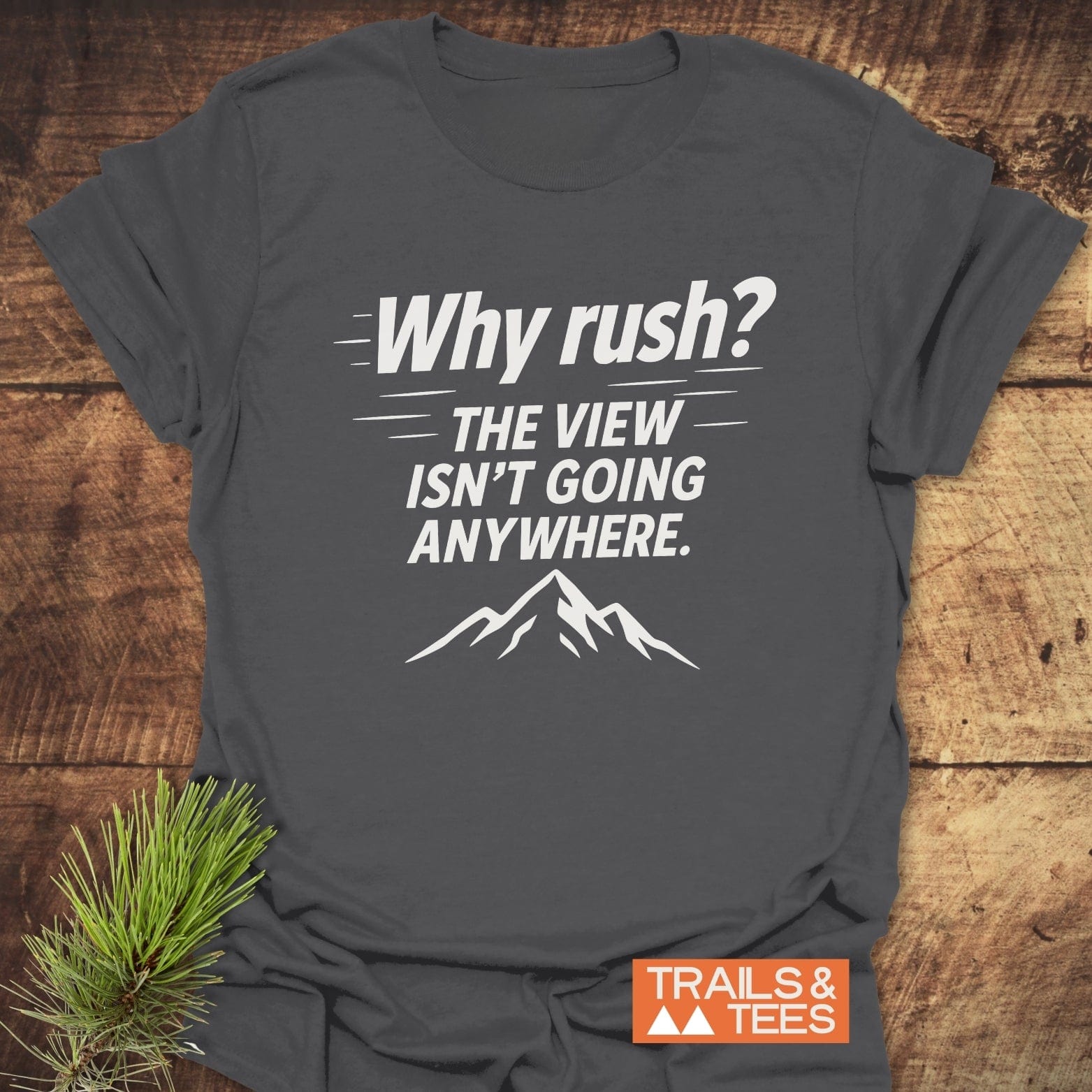 Why Rush? Slow Hikers T-Shirt on wooden surface, featuring mountain silhouette. Soft, durable cotton, pre-shrunk, no side seams for comfort. Trails & Tees logo visible.