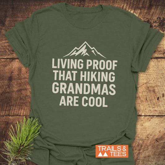 Living Proof Grandma T-Shirt in olive green, featuring a mountain silhouette and slogan, on wooden surface with pine branch and Trails & Tees logo visible.