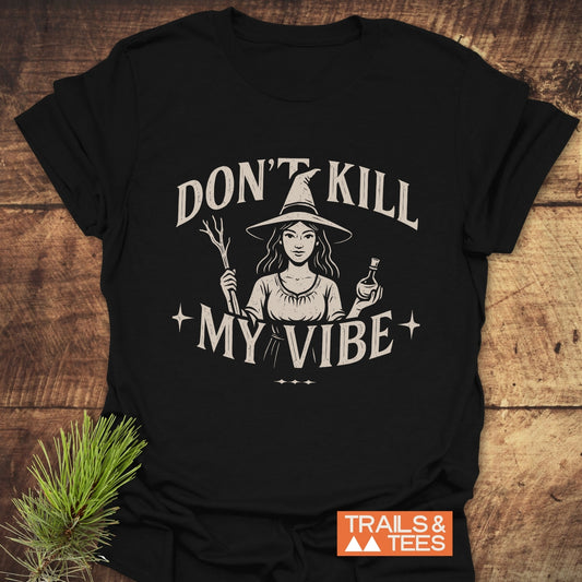 Don't Kill My Vibe Wood Witch T-Shirt featuring a witch illustration, slogan, and Trails & Tees logo, laid on a wooden surface with a pine sprig.