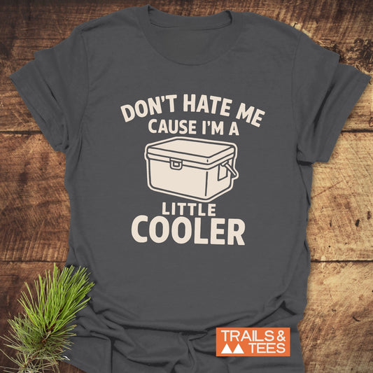 A Little Cooler Camping T-Shirt featuring a cooler graphic and playful text, laid on wood with a pine sprig, embodying outdoor adventure spirit.