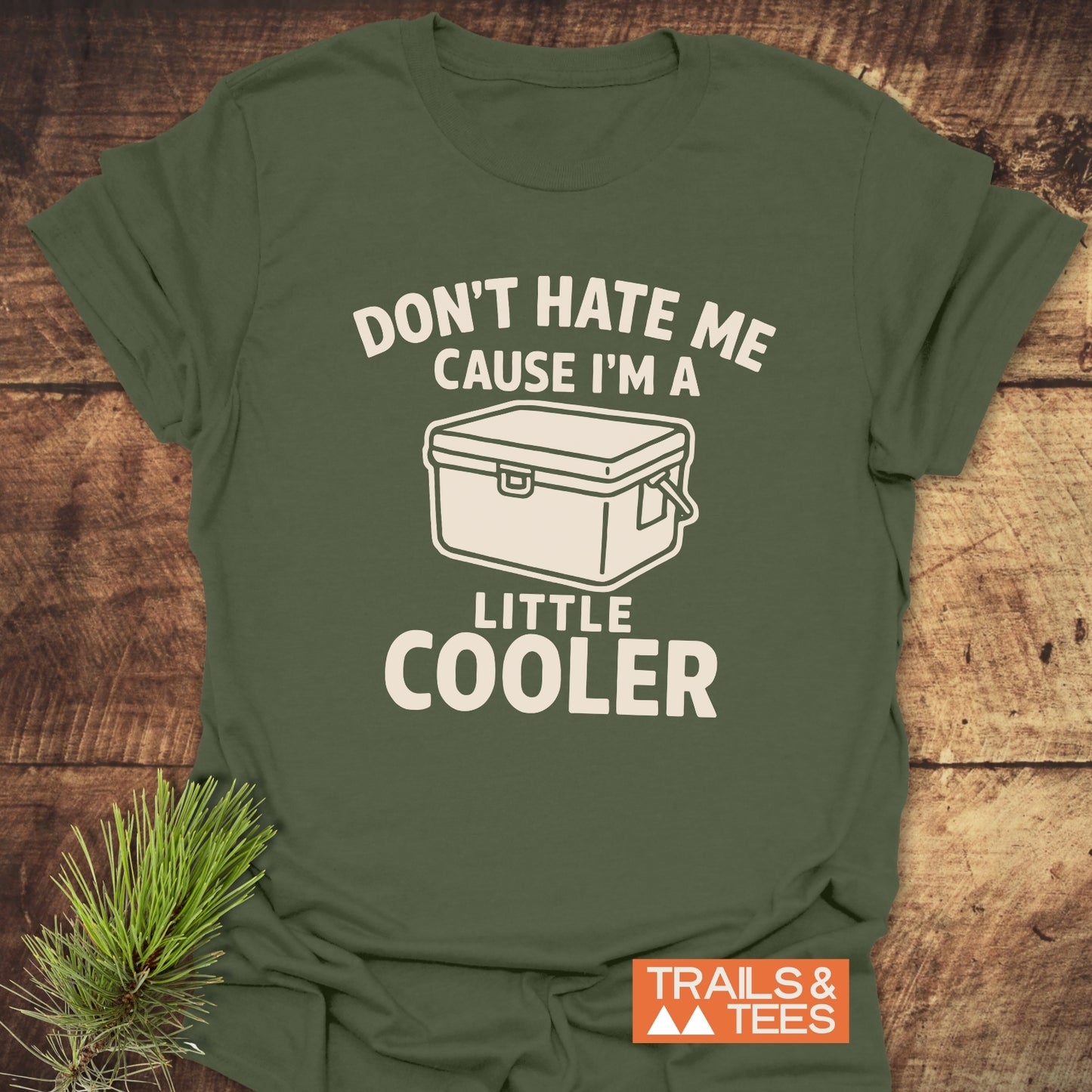 A Little Cooler Camping T-Shirt featuring a cooler graphic and slogan, laid flat with a pine sprig, highlighting its soft, adventure-ready cotton design.