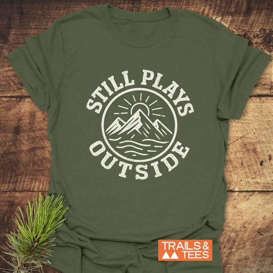 Still Plays Outside T-Shirt with mountain-sun icon, pine sprig, and Trails & Tees logo; crafted for comfort and adventure with durable, eco-friendly design.