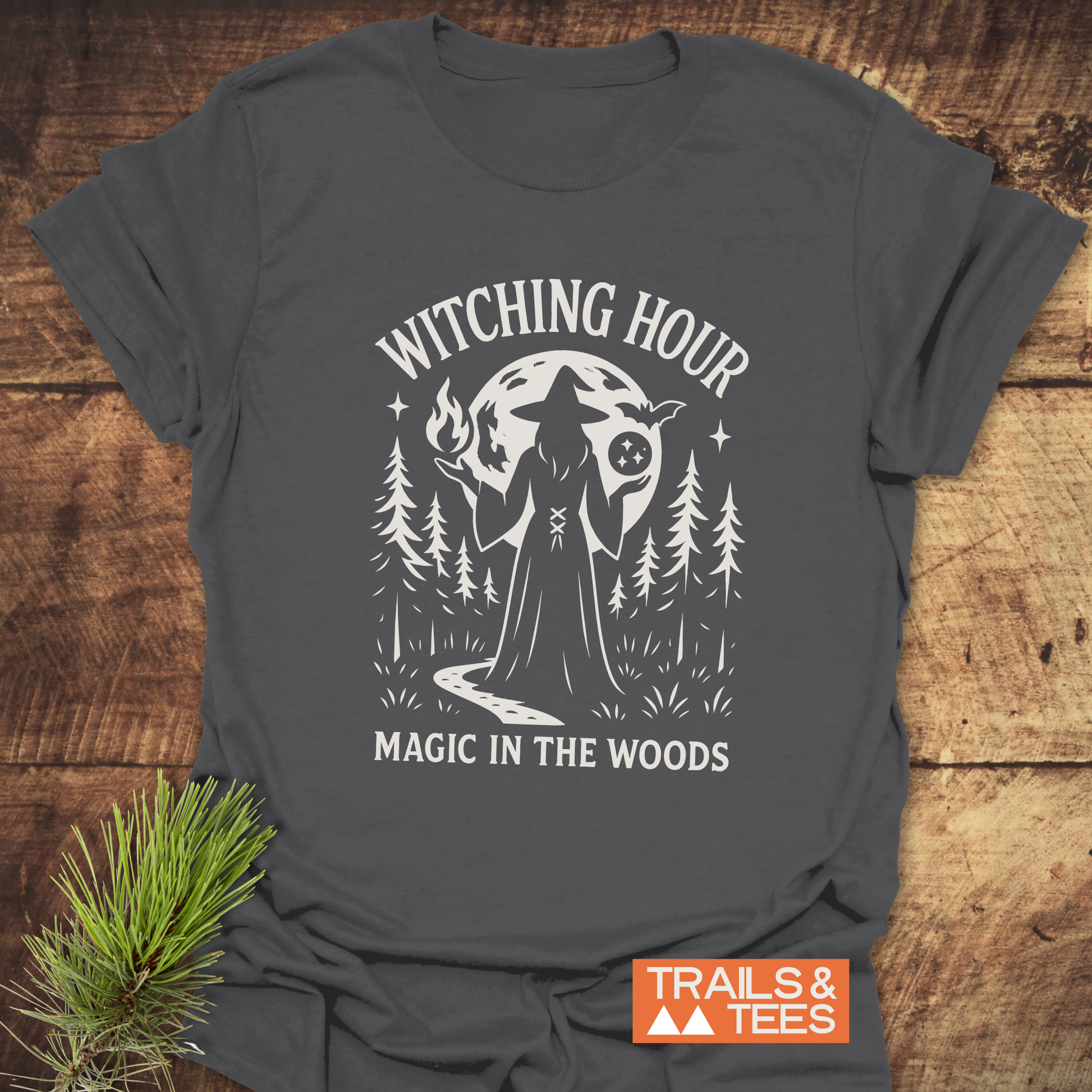 Witching Hour T-Shirt featuring a witch silhouette with pine trees, moon, and Magic in the Woods text. Durable, soft cotton, perfect for outdoor adventures.