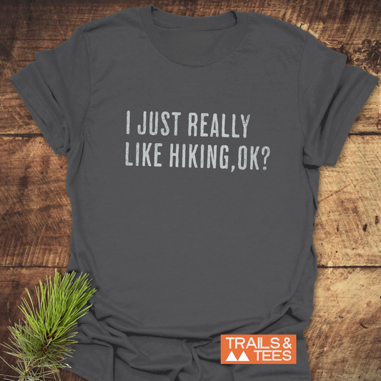 Dark-gray T-shirt reading “I JUST REALLY LIKE HIKING, OK?” with “TRAILS & TEES” logo, laid on wood, highlighting comfort and adventure-ready design.