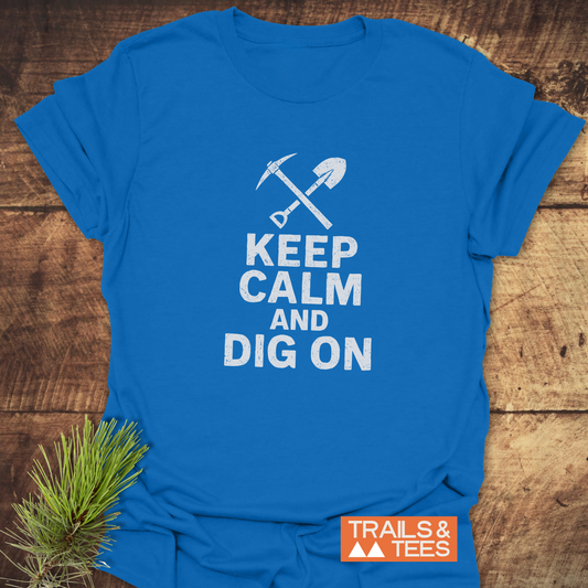 Keep Calm Rock Lover T-Shirt featuring a white pickaxe and shovel graphic with slogan, laid on wood with a pine branch, showcasing Trails & Tees logo.