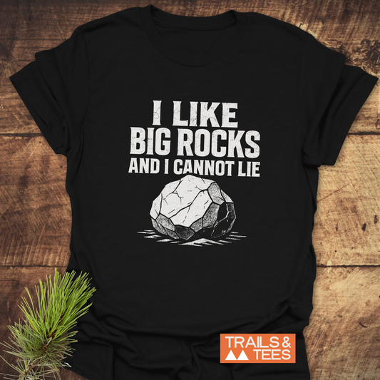 I Like Big Rocks Funny T-Shirt on wooden surface, features white slogan and rock design. Showcases Trails & Tees logo, emphasizing outdoor comfort and durability.
