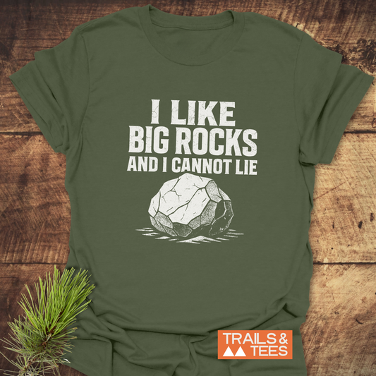 Olive-green I Like Big Rocks Funny T-Shirt with bold slogan and rock illustration, featuring a TRAILS & TEES logo, laid on a wooden surface.