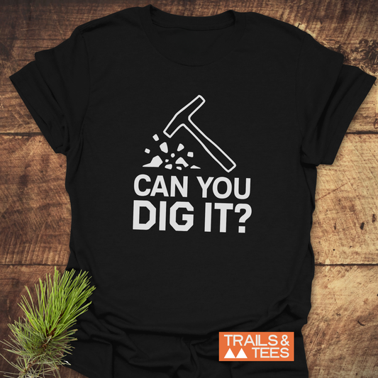 Black Can You Dig It? Rock Lovers T-Shirt with rock-hammer graphic, showcasing Trails & Tees logo, laid on wood with pine sprig.