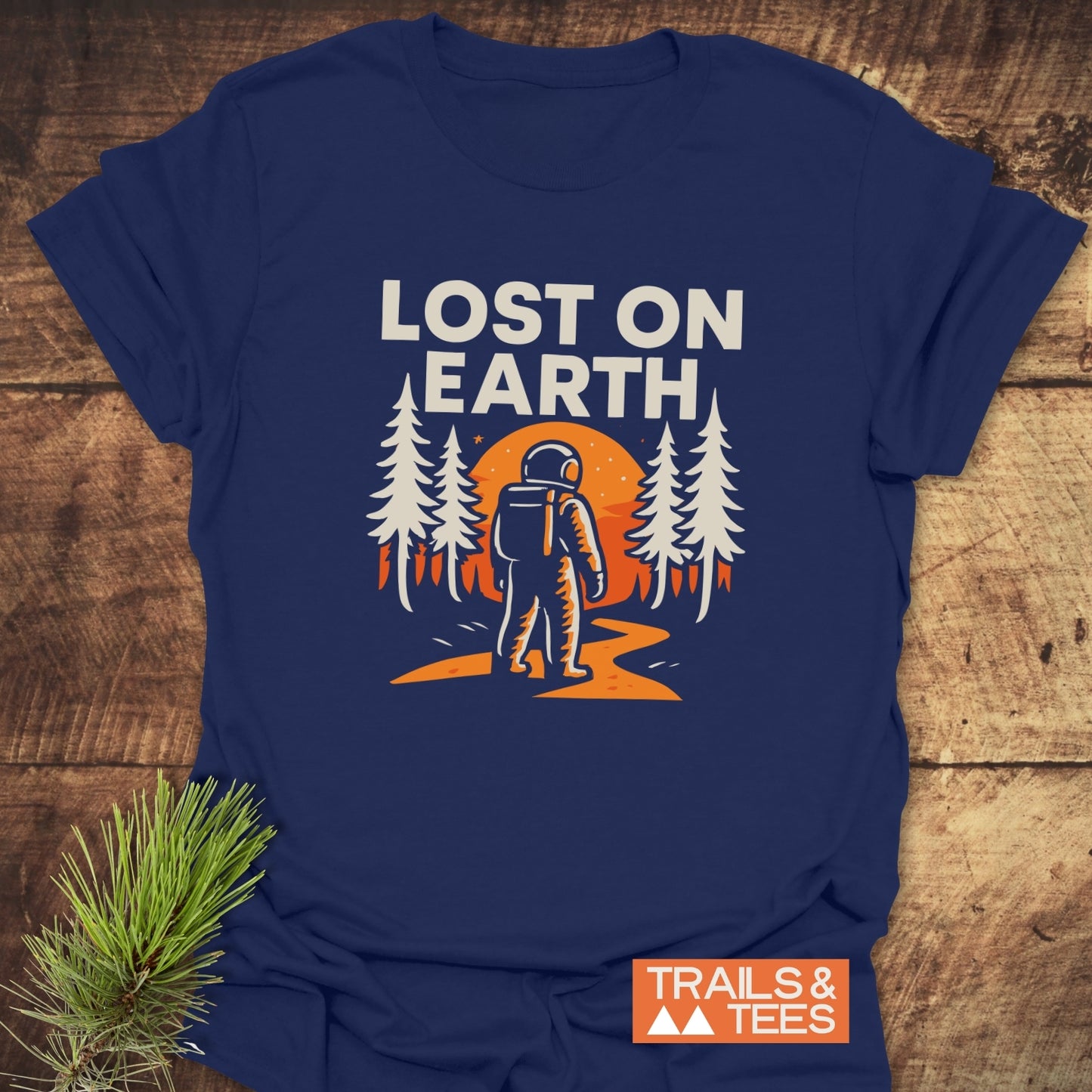 Lost On Earth T-Shirt featuring an astronaut in a pine forest with orange sun/moon, styled on wood with pine sprig, and Trails & Tees branding.