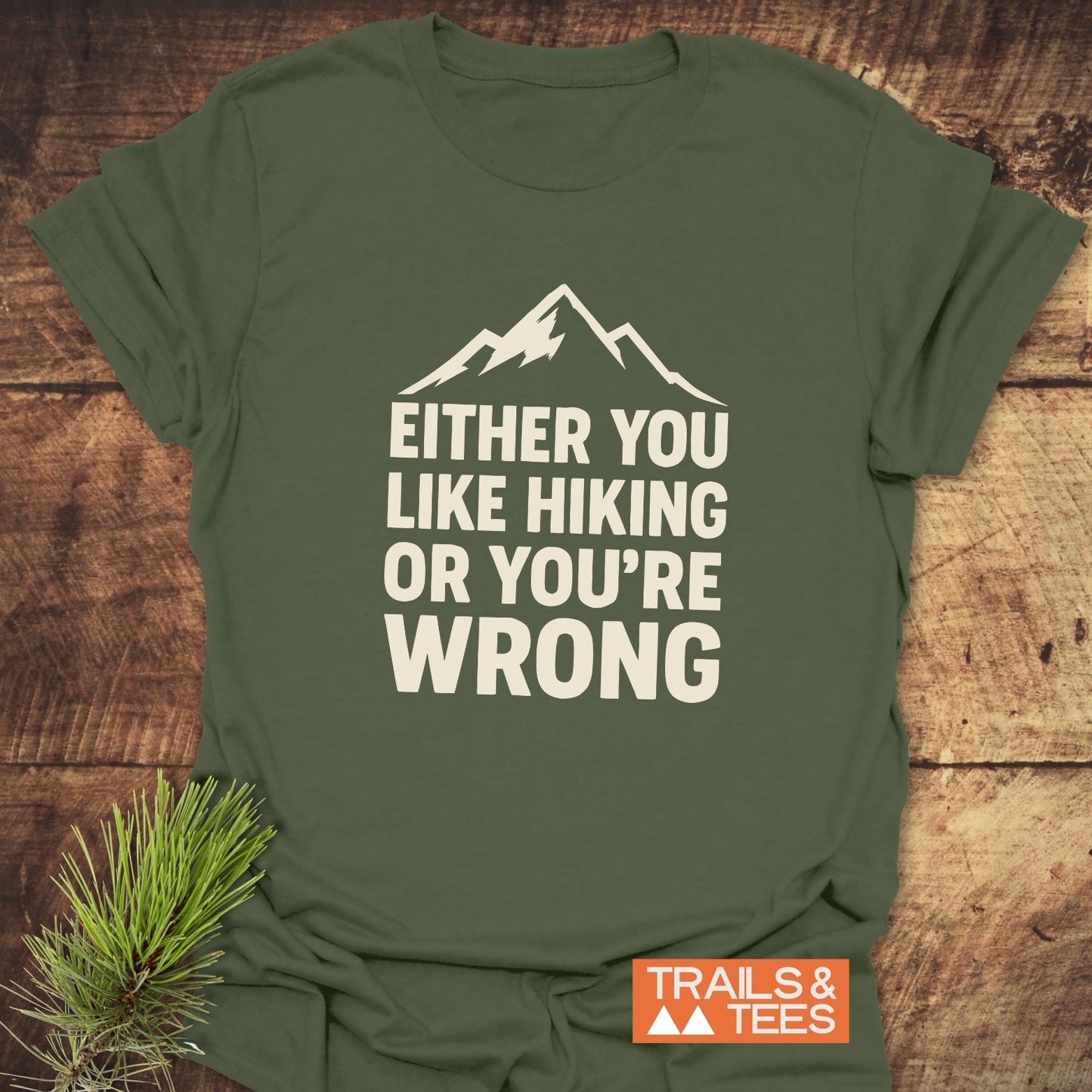 Army-green T-shirt with mountain graphic and text “EITHER YOU LIKE HIKING OR YOU’RE WRONG,” laid flat on wood surface, next to pine needles.