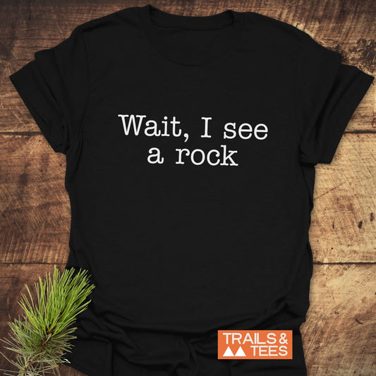 Wait, I See A Rock T-Shirt featuring white typewriter-style text on black, laid flat with pine sprig; includes Trails & Tees logo, crafted for outdoor adventures.