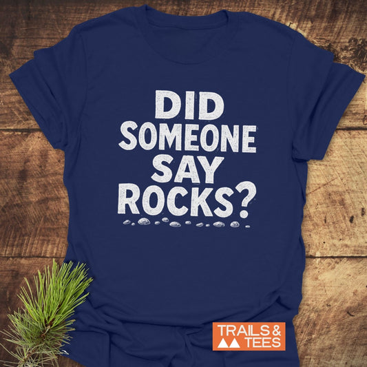 Did Someone Say Rocks? T-Shirt with white distressed lettering and pebble illustrations, branded with Trails & Tees logo, showcasing outdoor-themed design on a navy-blue background.