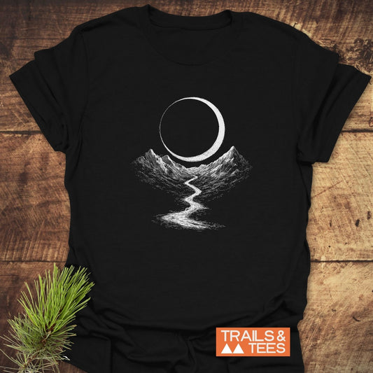 Eclipse Trail T-Shirt: Black tee with a crescent and mountain design, showcasing outdoors spirit. Includes TRAILS & TEES logo, staged with pine needles.