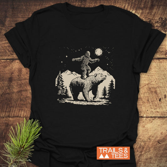Bigfoot And Bear Funny T-Shirt featuring a whimsical forest scene design on a black tee, displayed with a pine branch, highlighting outdoor adventure themes.