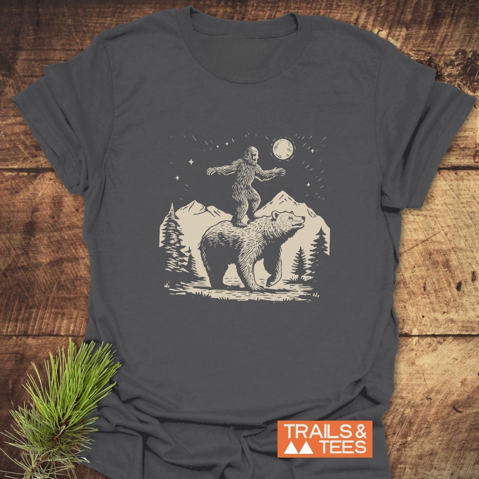 Bigfoot And Bear Funny T-Shirt on wood surface, showcasing Sasquatch balancing on a bear, with mountain scenery. Features soft, durable cotton for outdoor enthusiasts.