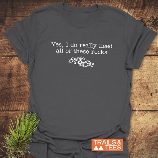 All These Rocks T-Shirt on wooden surface, featuring white text and stone sketch. Includes TRAILS & TEES logo. Made from soft, ring-spun cotton for outdoor enthusiasts.