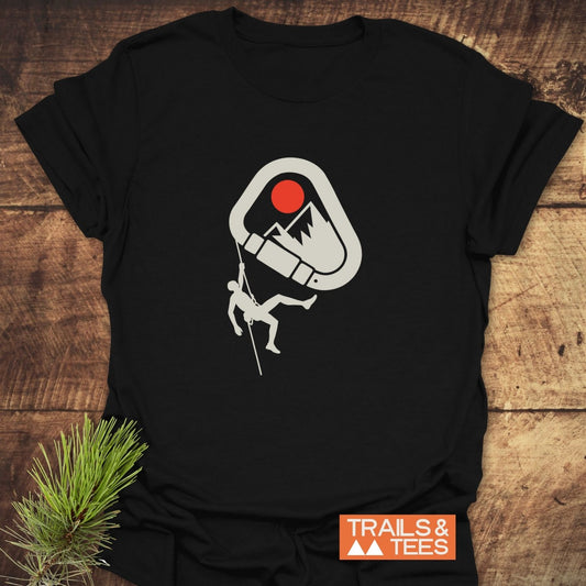587 Mountain Climber Silhouette T-Shirt featuring a carabiner with a mountain and climber graphic, laid on wood with a pine sprig, TRAILS & TEES logo.