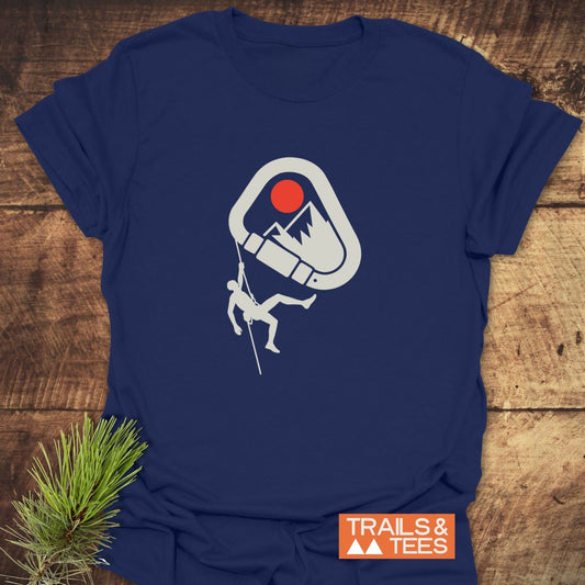 587 Mountain Climber Silhouette T-Shirt features a climber graphic on navy-blue cotton, symbolizing outdoor adventure. Laid on wood, with TRAILS & TEES logo nearby.