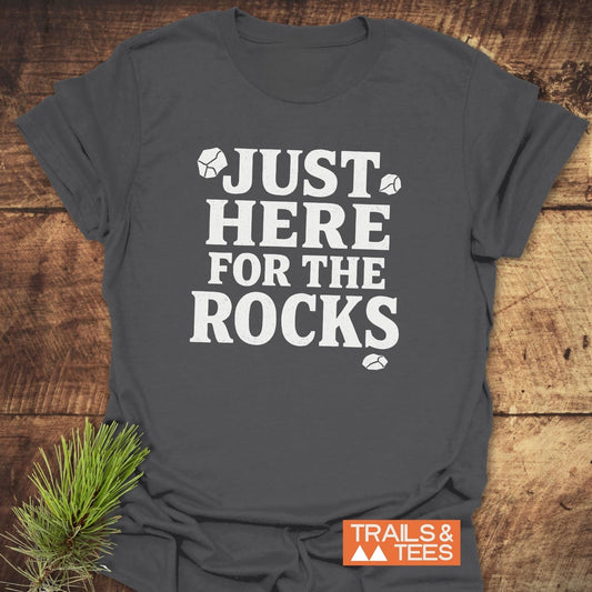 Just Here For The Rocks T-Shirt on rustic wood, with white text and rock icons. Orange “TRAILS & TEES” logo. Pine sprig accent.