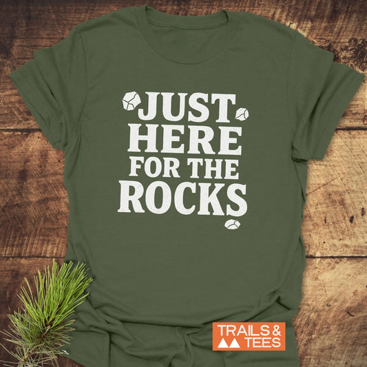 Just Here For The Rocks T-Shirt flat-lay, featuring bold white print on olive-green fabric, resting on wood with a pine sprig beside it.