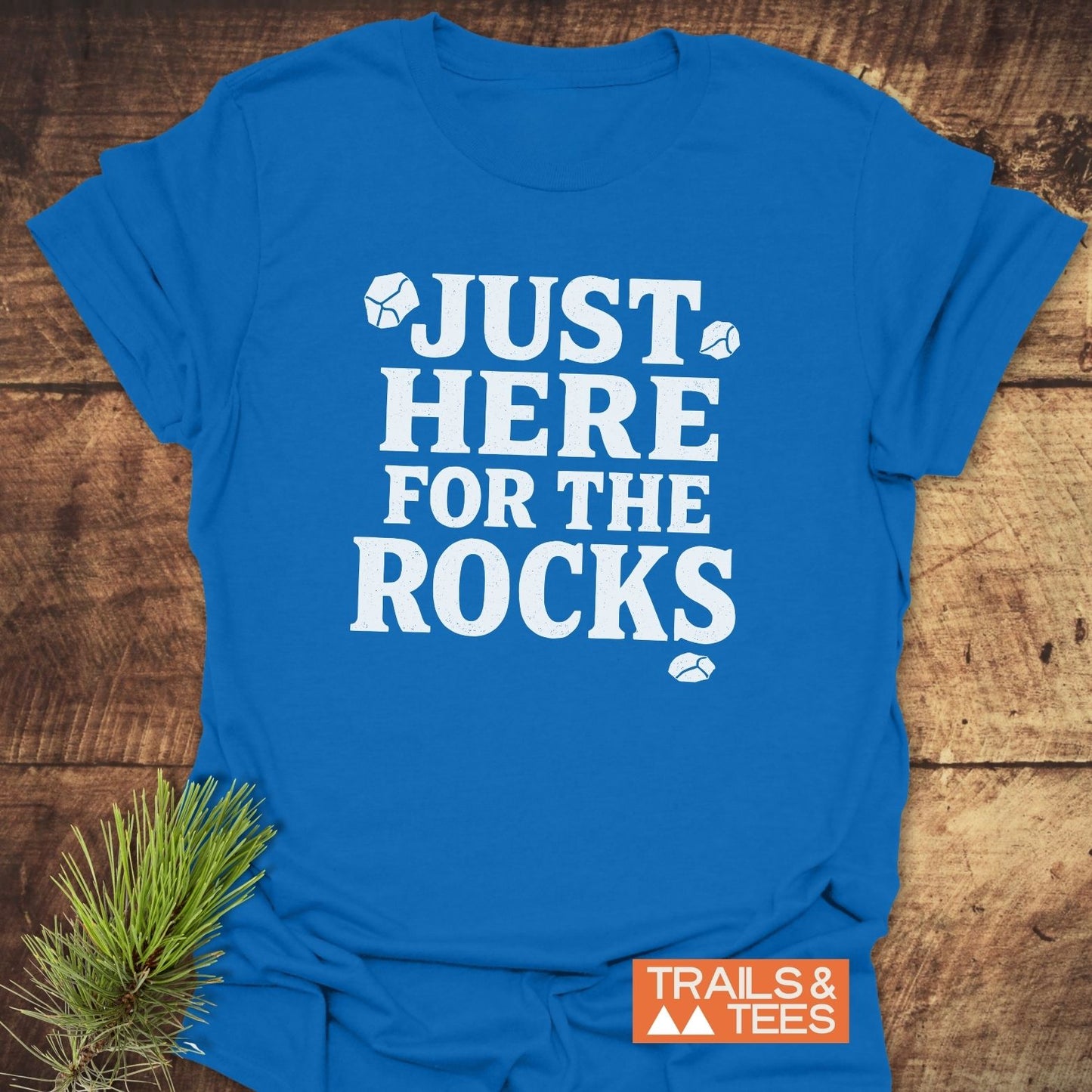 Just Here For The Rocks T-Shirt on wooden surface, features bold slogan with rock icons, orange Trails & Tees logo, and pine sprig styling.