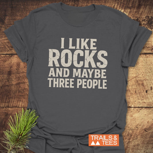 Charcoal I Like Rocks T-Shirt on wood background with pine sprig, featuring off-white slogan, durable design, and small TRAILS & TEES logo nearby.