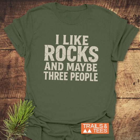 I Like Rocks T-Shirt with I LIKE ROCKS AND MAYBE THREE PEOPLE print, featuring Trails & Tees logo, displayed on wood with pine sprig.
