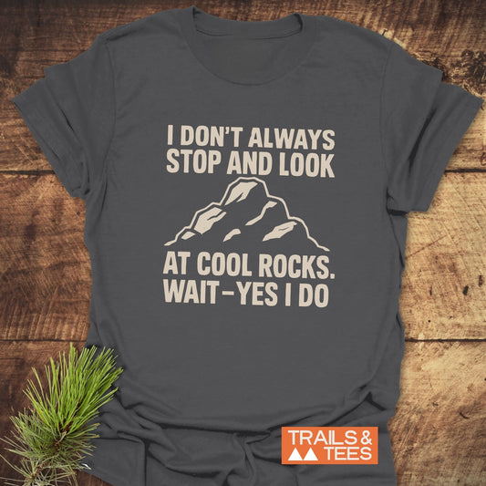Dark-grey T-shirt with mountain peak graphic and the text I DON'T ALWAYS STOP AND LOOK AT COOL ROCKS. WAIT – YES I DO. Features TRAILS & TEES logo.