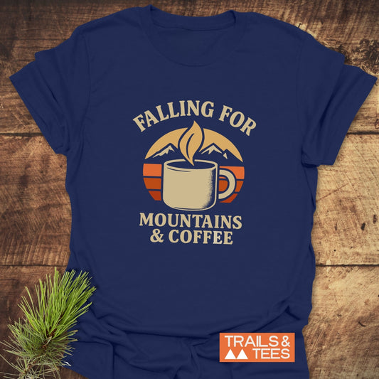 Falling For T-Shirt with coffee mug and mountain graphic, slogan “FALLING FOR MOUNTAINS & COFFEE.” Laid on wood surface, Trails & Tees logo visible.