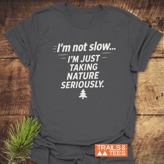 Charcoal-gray I'm Not Slow... Funny T-Shirt with white slogan and pine icon, laid flat with a pine sprig, showcasing Trails & Tees logo.