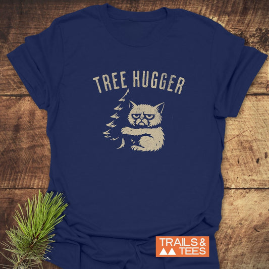 Tree Hugger Funny T-Shirt featuring a grumpy cat hugging a tree graphic, displayed on wooden surface with pine sprig, ideal for outdoor enthusiasts.