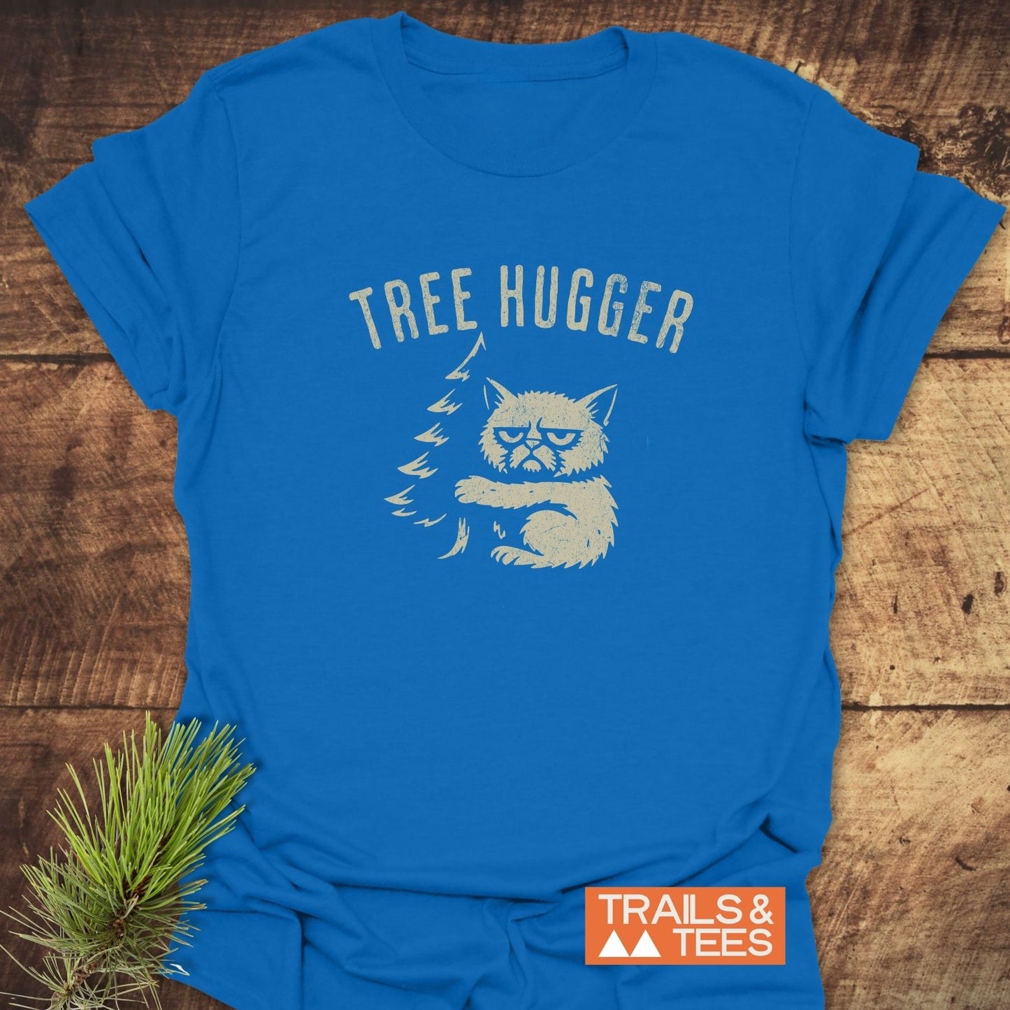 Tree Hugger Funny T-Shirt with a distressed print of a cat hugging a pine tree, showcasing the “TRAILS & TEES” logo, resting on a wooden surface.