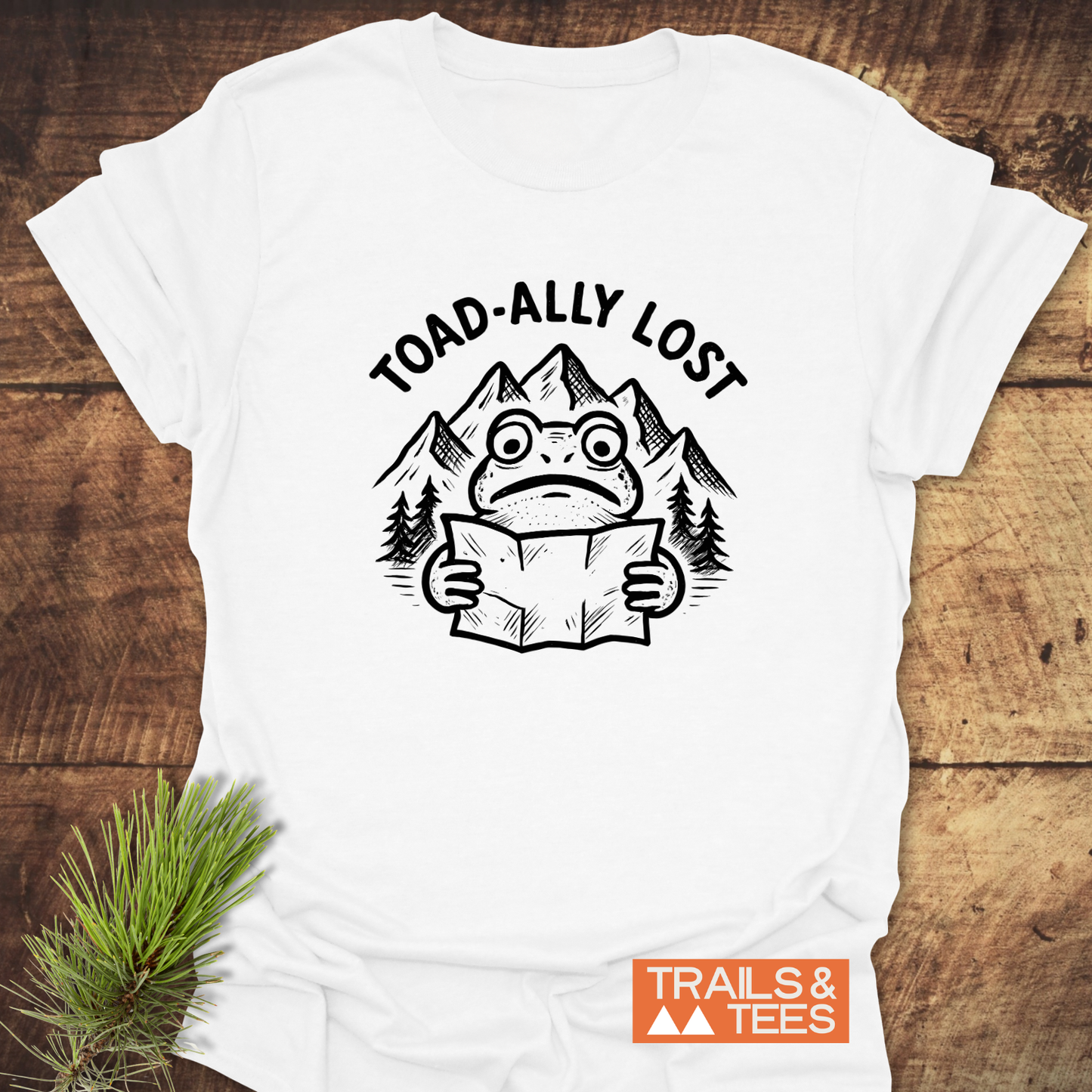 Toad-ally Lost T-Shirt featuring a toad with a map, pine trees, and mountains. Crafted from soft cotton, with durable print and a Trails & Tees logo.