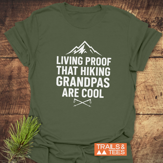 Forest-green 606 Living Proof Grandpa T-Shirt with mountain graphic and slogan, featuring DTF prints and crossed hiking poles. Laid out on a wooden surface.