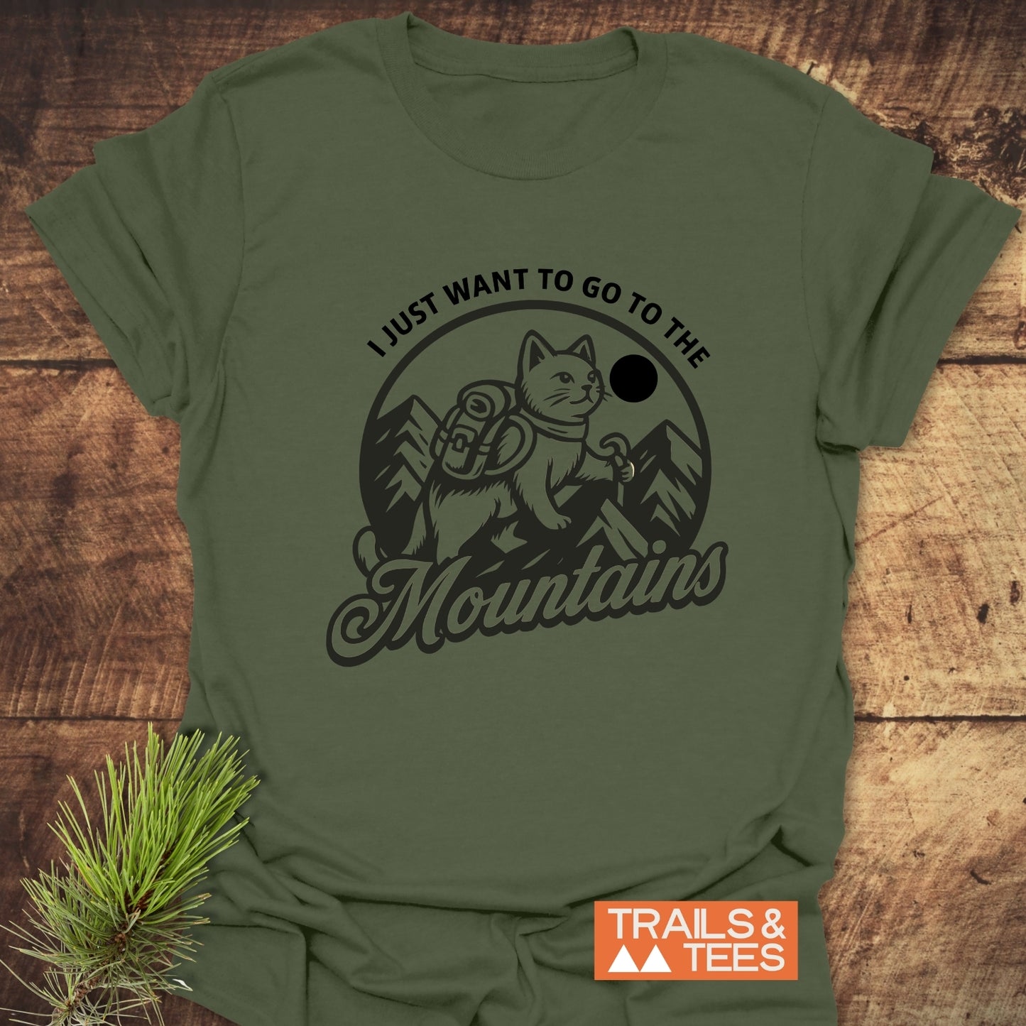 Mountain Kitty T-Shirt featuring a cat hiking graphic, crafted from soft ring-spun cotton for comfort and durability, laid flat with a pine sprig beside it.