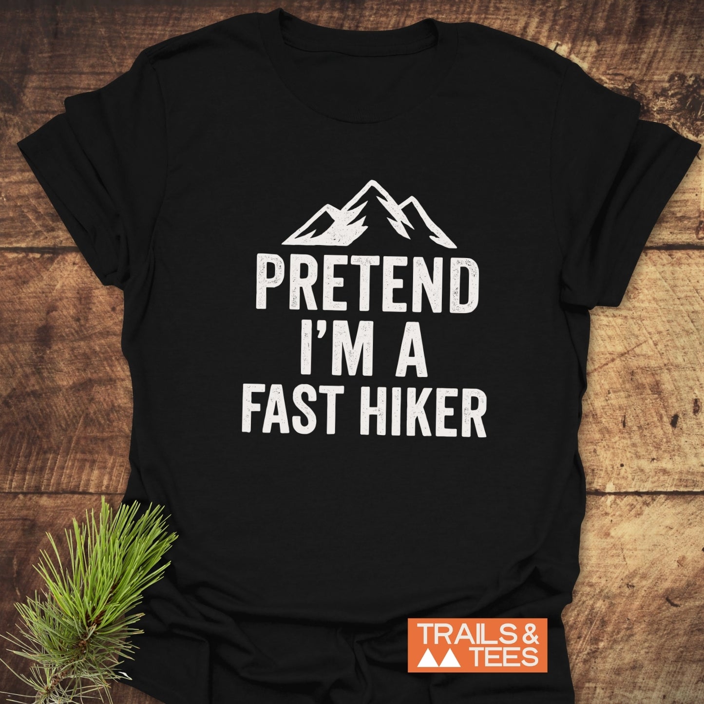 Black 'Pretend I'm A Fast Hiker' T-shirt with mountain graphic, laid on wood with pine sprig, emphasizes comfort and outdoor spirit from Trails & Tees.