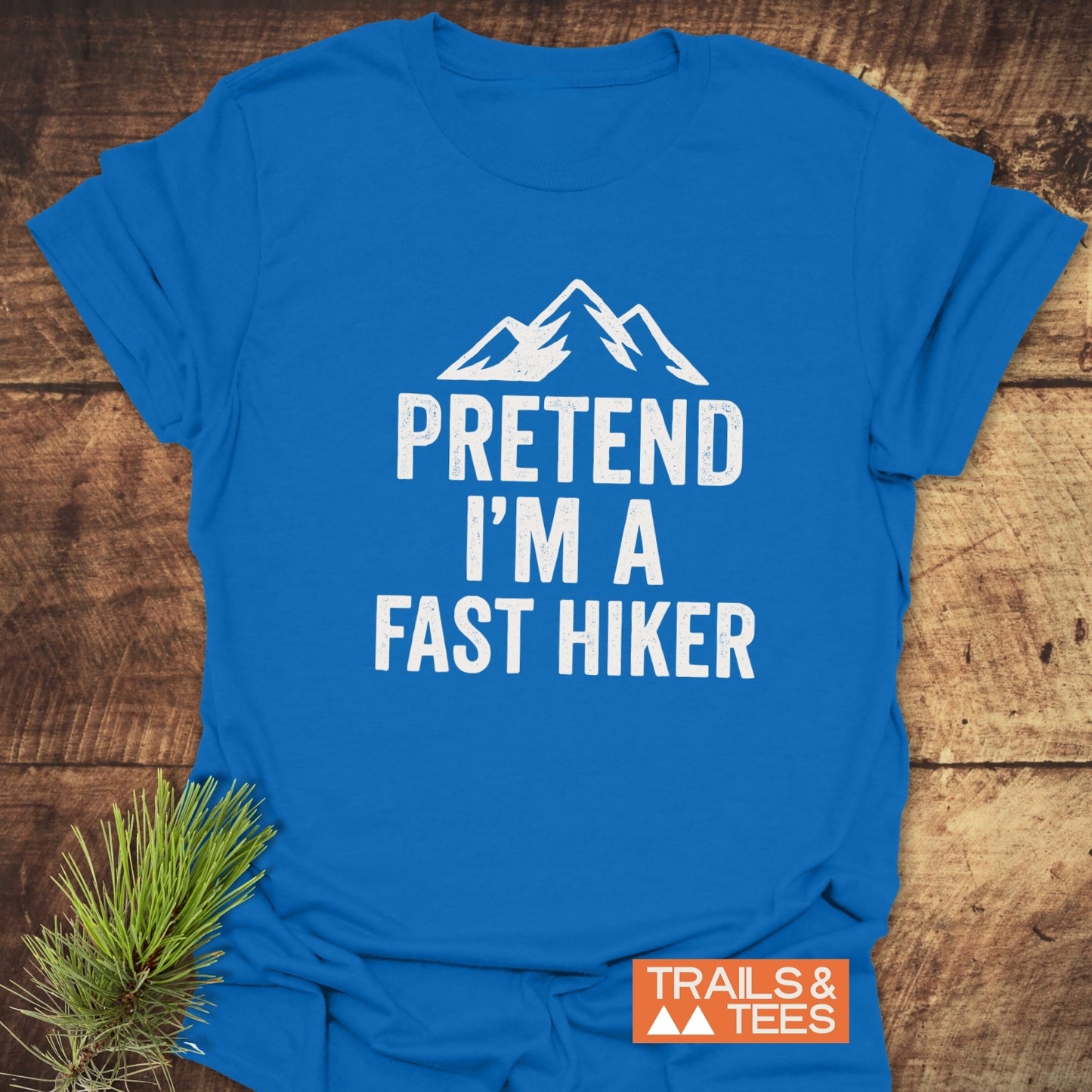 Pretend I'm A Fast Hiker T-Shirt with mountain graphic, laid on wood, showcases durable, soft cotton for outdoor enthusiasts. Pine sprig adds a nature touch.