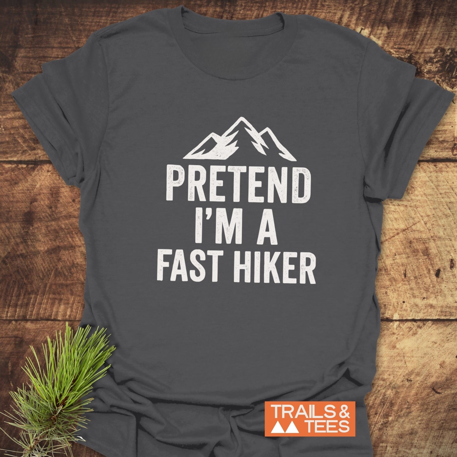 Charcoal-gray Pretend I'm A Fast Hiker T-shirt with mountain design on rustic surface, featuring Trails & Tees logo and pine twig styling.