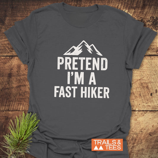 Charcoal-gray Pretend I'm A Fast Hiker T-shirt with mountain design on rustic surface, featuring Trails & Tees logo and pine twig styling.