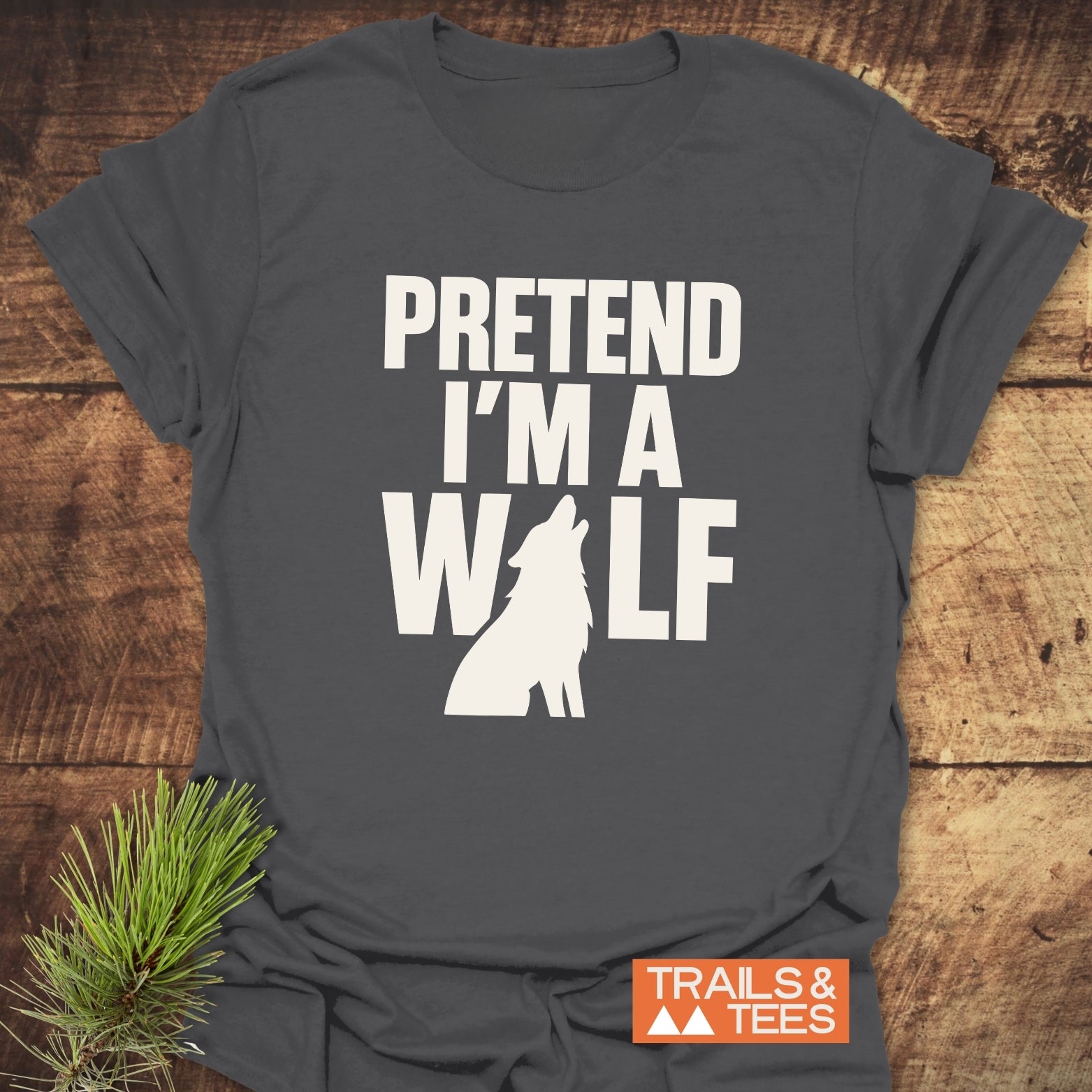 Pretend I'm A Wolf T-Shirt with bold white text and wolf silhouette, featuring soft, durable cotton and small Trails & Tees logo, laid on wooden surface.