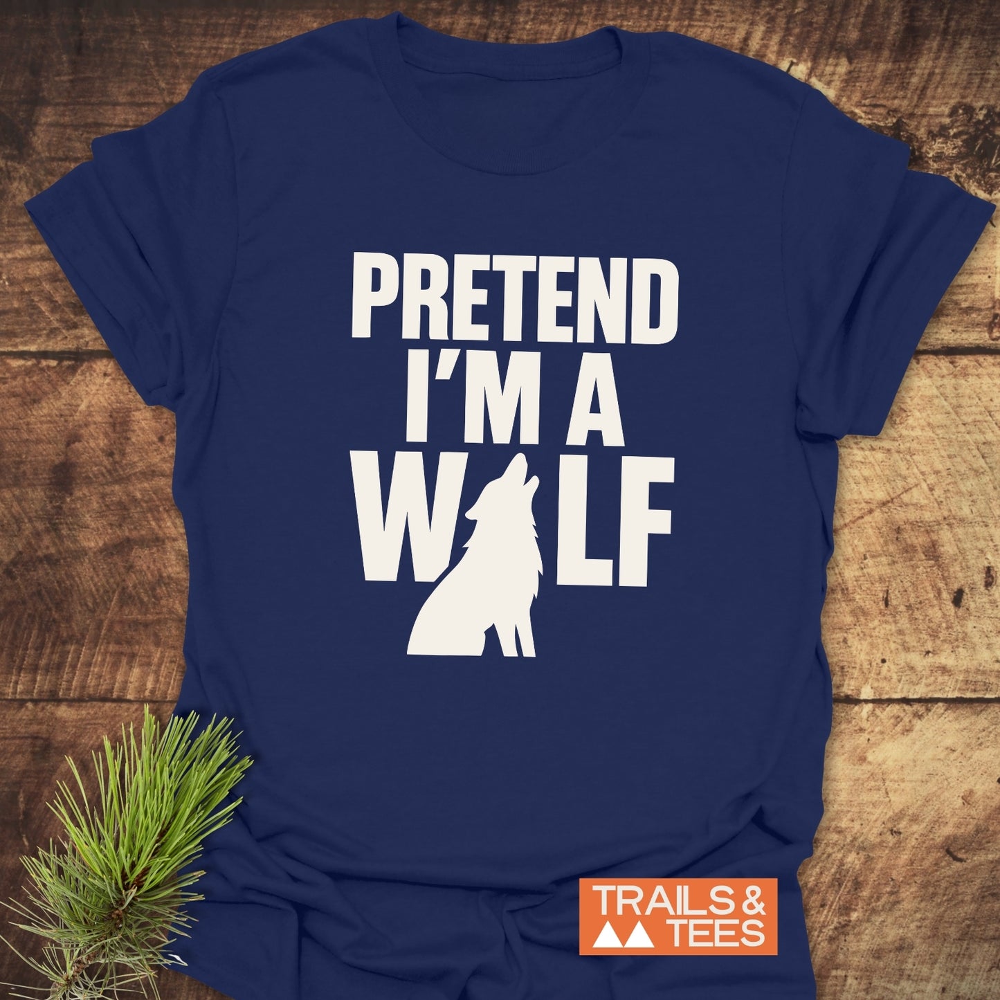 Navy blue Pretend I'm A Wolf T-shirt with a wolf silhouette, on rustic wood, featuring Trails & Tees logo. Pine sprig adds a nature vibe.