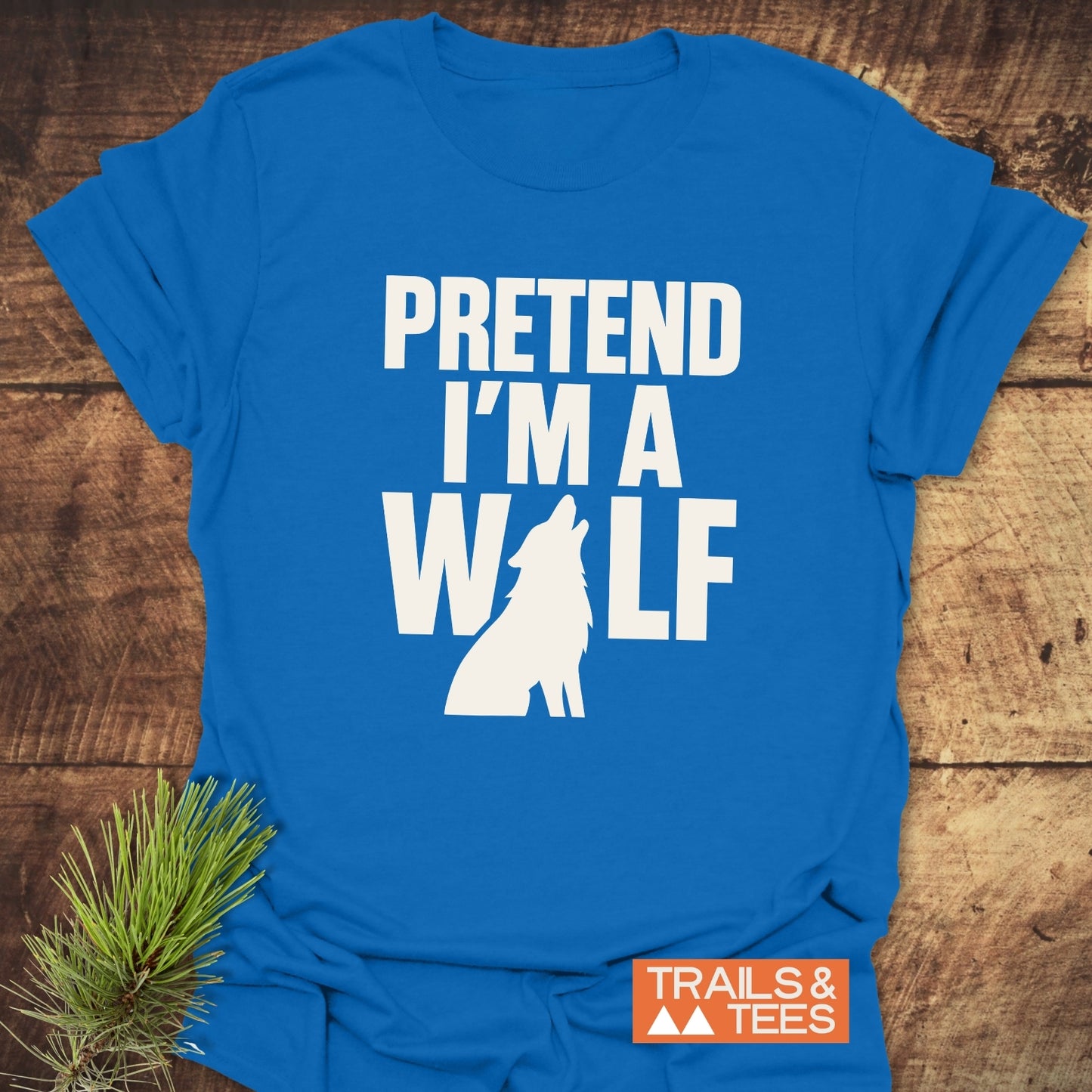 Pretend I'm A Wolf T-Shirt featuring bold white text and wolf silhouette, laid on wood. Soft, lightweight cotton with durable print and Trails & Tees logo.