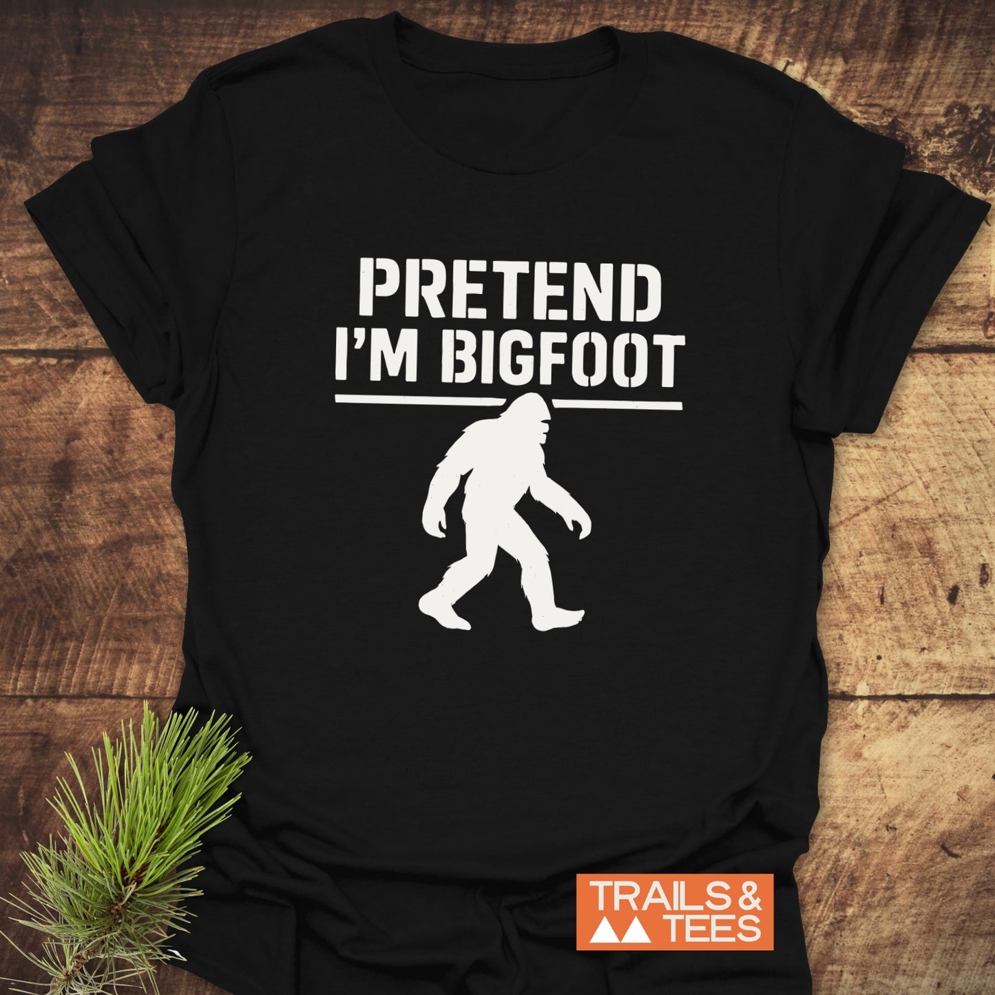 Black 'Pretend I'm Bigfoot' T-shirt with white text and Bigfoot silhouette, laid flat with orange 'TRAILS & TEES' logo, suggesting an outdoor theme.